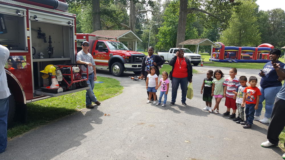 Two men showing adults and children a firetruck at a park
