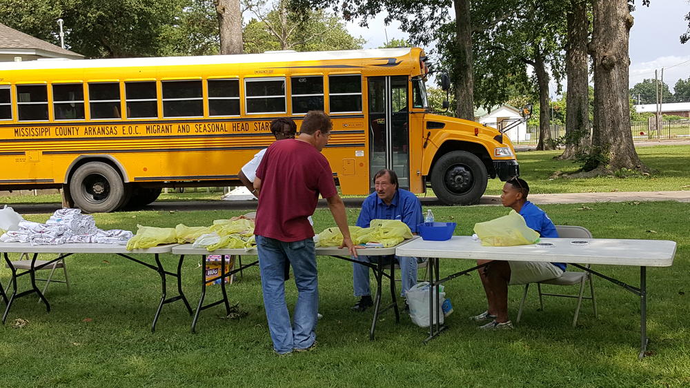 Four adults talking at tables with stacks of shirts and a school bus in the back