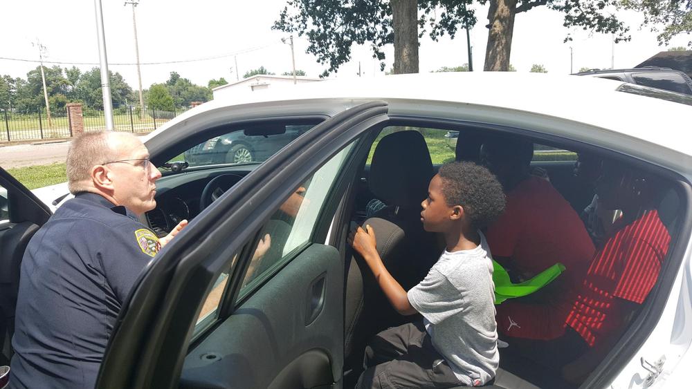 Adults and children sitting in the back of a police car while the officer talks to them